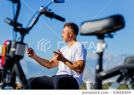 old man with gray beard sitting on the view poing with mountain and beach next to electro bike sunny day using smartphone old man with gray beard sitting on the view poing with mountain and beach next to electro bike sunny day using smartphone 93668904