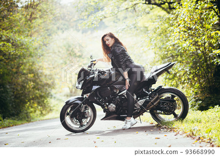 Portrait of a beautiful young woman sitting on a motorcycle, riding a motorcycle in black leather clothes on nature, street, forest, in sunny weather, in full growth 93668990