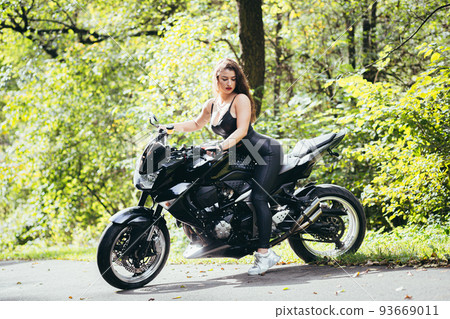 Portrait of a beautiful young woman sitting on a motorcycle, riding a motorcycle in black leather clothes on nature, street, forest, in sunny weather, in full growth 93669011