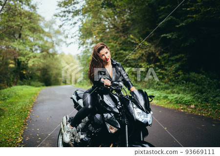Portrait of a beautiful young woman sitting on a motorcycle, riding a motorcycle in black leather clothes on nature, street, forest, in sunny weather, in full growth 93669111