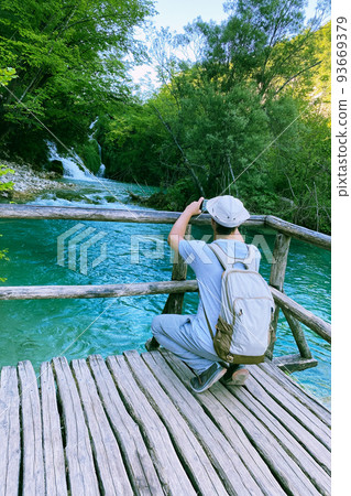 Tourist takes pictures of the river on a smartphone. Summer landscapes along lake in journey. Wooden vintage bridge walkway. 93669379
