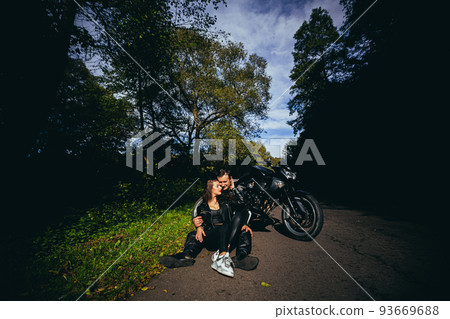 Handsome young man and beautiful young girl, couple sitting near motorcycle in black leather clothes, hugging, in nature, outdoors Handsome young man and beautiful young girl, couple sitting near motorcycle in black leather clothes, hugging, in nature, outdoors 93669688