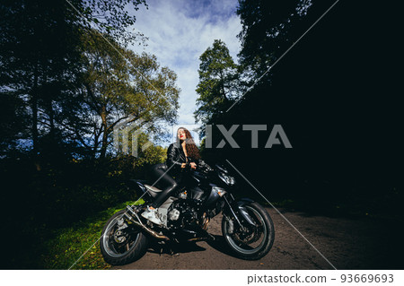 Portrait of a beautiful young woman sitting on a motorcycle, riding a motorcycle in black leather clothes on nature, street, forest, in sunny weather, in full growth 93669693