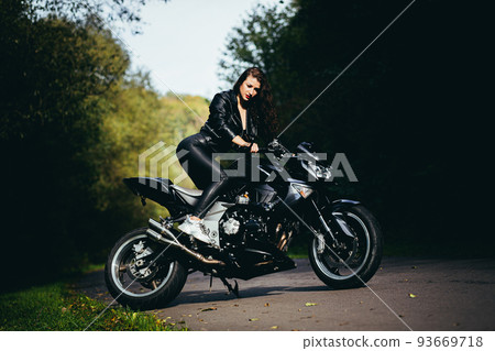 Portrait of a beautiful young woman sitting on a motorcycle, riding a motorcycle in black leather clothes on nature, street, forest, in sunny weather, in full growth 93669718