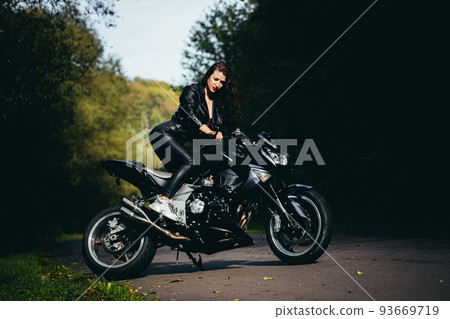Portrait of a beautiful young woman sitting on a motorcycle, riding a motorcycle in black leather clothes on nature, street, forest, in sunny weather, in full growth 93669719