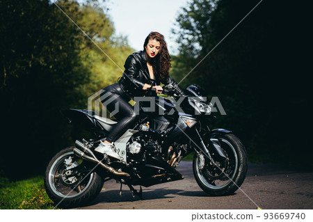Portrait of a beautiful young woman sitting on a motorcycle, riding a motorcycle in black leather clothes on nature, street, forest, in sunny weather, in full growth 93669740