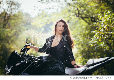 Portrait of a beautiful young woman on a motorcycle in black leather clothes on the nature, street, forest, in sunny weather, close up, looking at the camera Portrait of a beautiful young woman on a motorcycle in black leather clothes on the nature, street, forest, in sunny weather, close up, looking at the camera 93669767