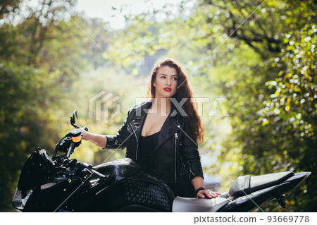 Portrait of a beautiful young woman on a motorcycle in black leather clothes on the nature, street, forest, in sunny weather, close up, looking at the camera Portrait of a beautiful young woman on a motorcycle in black leather clothes on the nature, street, forest, in sunny weather, close up, looking at the camera 93669778