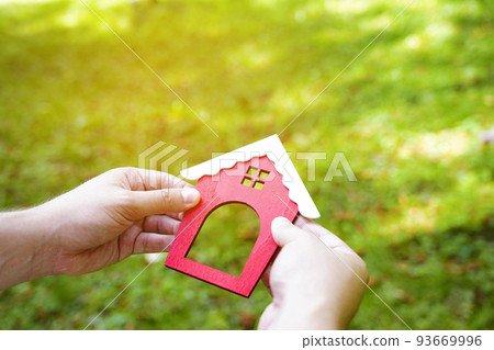 Miniature wooden house in the hands of a man outdoors. Real estate concept. Eco-friendly home. Buying a housing outside the city. Red roof. Selective focus Miniature wooden house in the hands of a man outdoors. Real estate concept. Eco-friendly home. Buying a housing outside the city. Red roof. Selective focus 93669996