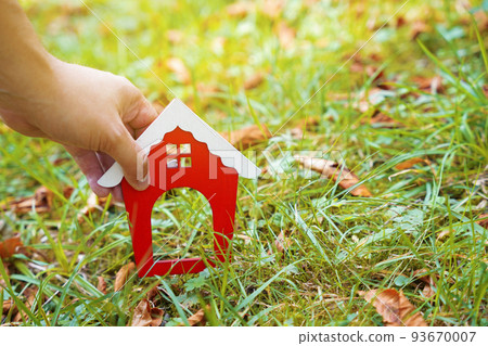 Miniature wooden house in the hands of a man outdoors. Real estate concept. Eco-friendly home. Buying a housing outside the city. Red roof. Selective focus 93670007