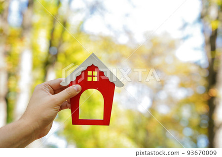 Miniature wooden house in the hands of a man outdoors. Real estate concept. Eco-friendly home. Buying a housing outside the city. Red roof. Selective focus 93670009