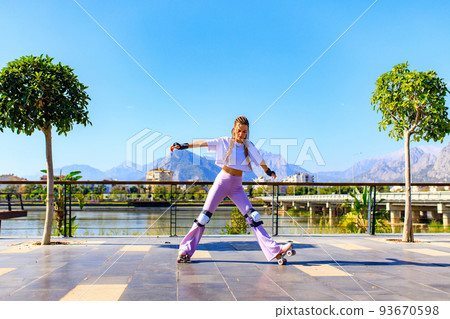 Portrait of beautiful blonde teenager girl with cool pigtails , white t-shirt and flared pink pants skating on rollers in park 93670598