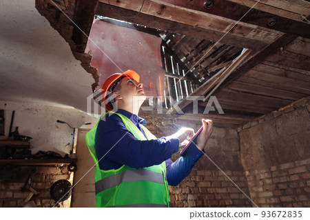 Building inspector. Man in a hard hat and a yellow reflective vest examines damaged structures and inspects the building. Damage assessment. Preparing for the repair or construction of a building Building inspector. Man in a hard hat and a yellow reflective vest examines damaged structures and inspects the building. Damage assessment. Preparing for the repair or construction of a building 93672835