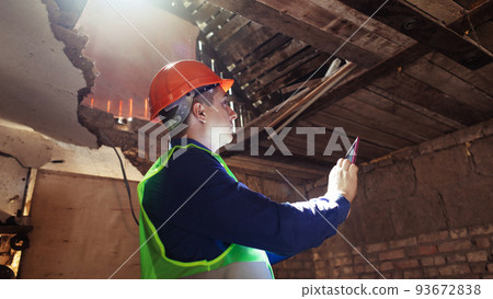 Building inspector. Man in a hard hat and a yellow reflective vest examines damaged structures and inspects the building. Damage assessment. Preparing for the repair or construction of a building Building inspector. Man in a hard hat and a yellow reflective vest examines damaged structures and inspects the building. Damage assessment. Preparing for the repair or construction of a building 93672838