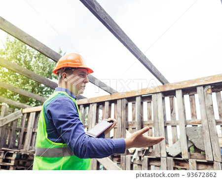 Building inspector. Man in a hard hat and a yellow reflective vest examines damaged structures and inspects the building. Damage assessment. Preparing for the repair or construction of a building Building inspector. Man in a hard hat and a yellow reflective vest examines damaged structures and inspects the building. Damage assessment. Preparing for the repair or construction of a building 93672862