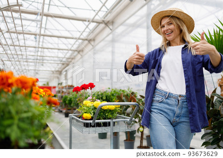 Pretty woman in garden center or greenhouse with shopping cart showing thumbs up rejoicing at purchased flowers 93673629
