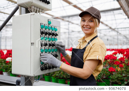 Pleasant woman in apron and cap presses the buttons on the  control panel of the automatic watering system for flowers in the greenhouse 93674078