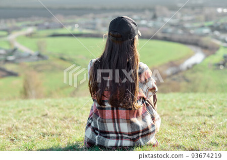 a girl sits with her back on a hill, enjoys a beautiful view a girl sits with her back on a hill, enjoys a beautiful view 93674219