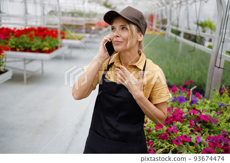 Pleasant woman working as a florist in a greenhouse talking to a smartphone with client about selling flowers 93674474