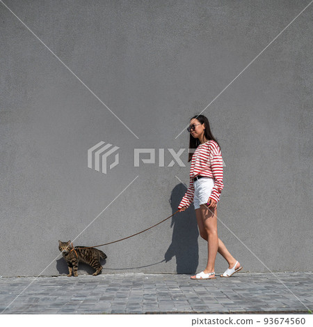 A young woman walks with a gray tabby cat on a leash against a gray wall.  93674560