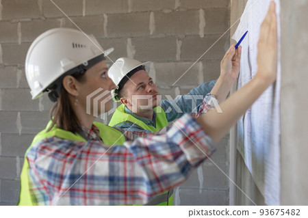 two engineers, a man and a woman in white helmets and protective vests, are standing in the room and holding a plan of the construction two engineers, a man and a woman in white helmets and protective vests, are standing in the room and holding a plan of the construction 93675482