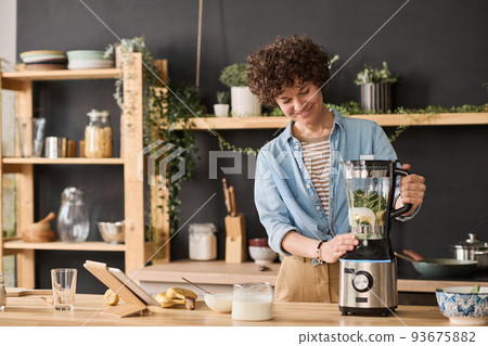 Young girl making fresh smoothie from fruits and milk in blender in the kitchen Young girl making fresh smoothie from fruits and milk in blender in the kitchen 93675882