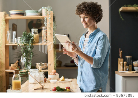 Young woman with curly hair reading recipe online using her digital tablet while cooking in kitchen Young woman with curly hair reading recipe online using her digital tablet while cooking in kitchen 93675893