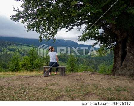 Female in casual clothes sits under large tree with forest background Female in casual clothes sits under large tree with forest background 93676317