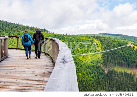 Couple of tourists with backpacks walking on the Sky walk in Dolni Morava with suspension Sky Bridge 721 on the background.  93676431