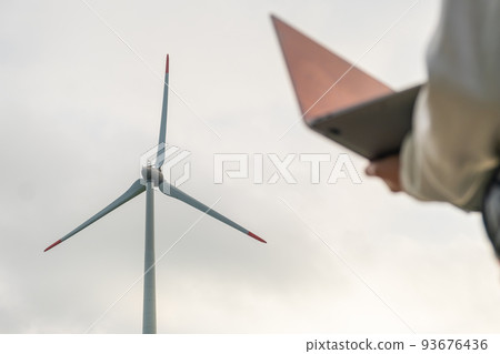 Close up woman uses a laptop to control the work of the windmill against the grey sky. Close up woman uses a laptop to control the work of the windmill against the grey sky. 93676436
