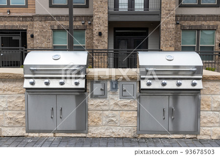 Two outdoor built in barbecue BBQ stainless steel grills at back yard of the residential complex with brick building on background Two outdoor built in barbecue BBQ stainless steel grills at back yard of the residential complex with brick building on background 93678503