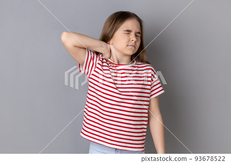 Portrait of unhealthy sick dark haired little girl wearing striped T-shirt standing massaging neck to relieve pain, muscle strain in back. Indoor studio shot isolated on gray background. Portrait of unhealthy sick dark haired little girl wearing striped T-shirt standing massaging neck to relieve pain, muscle strain in back. Indoor studio shot isolated on gray background. 93678522
