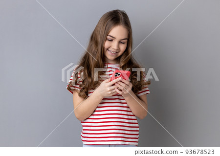 Portrait of smiling little girl wearing striped T-shirt using smart phone, looking at device screen, writing post for her kid's blog. Indoor studio shot isolated on gray background. Portrait of smiling little girl wearing striped T-shirt using smart phone, looking at device screen, writing post for her kid's blog. Indoor studio shot isolated on gray background. 93678523