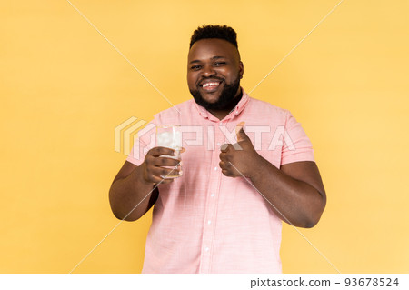 Portrait of satisfied smiling happy man wearing pink shirt feels thirsty, holding glass of cold fresh water with ice, showing thumb up. Indoor studio shot isolated on yellow background. Portrait of satisfied smiling happy man wearing pink shirt feels thirsty, holding glass of cold fresh water with ice, showing thumb up. Indoor studio shot isolated on yellow background. 93678524