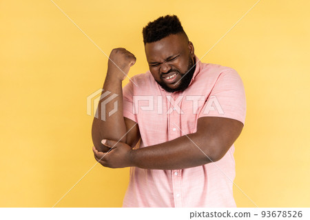 Portrait of man wearing pink shirt massaging aching elbow, frowning from acute pain, strain and strain problem, trapped nerves, rheumatoid arthritis. Indoor studio shot isolated on yellow background. Portrait of man wearing pink shirt massaging aching elbow, frowning from acute pain, strain and strain problem, trapped nerves, rheumatoid arthritis. Indoor studio shot isolated on yellow background. 93678526
