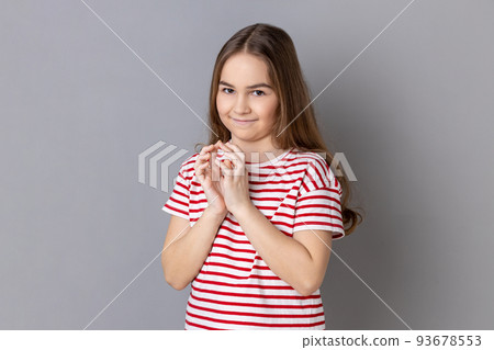 Portrait of cunning little girl wearing striped T-shirt standing looks at camera, planning something cunful, pranking somebody. Indoor studio shot isolated on gray background. Portrait of cunning little girl wearing striped T-shirt standing looks at camera, planning something cunful, pranking somebody. Indoor studio shot isolated on gray background. 93678553