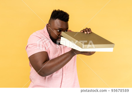 Portrait of curious attentive man wearing pink shirt holding present box in hands and looking inside with serious expression and big eyes. Indoor studio shot isolated on yellow background. 93678589