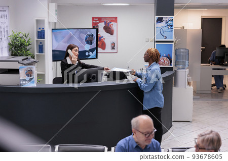 Receptionist using landline phone at reception desk, giving report papers to patient to fill in before medical checkup appointment. Worker helping woman with consultation forms, cord telephone. Receptionist using landline phone at reception desk, giving report papers to patient to fill in before medical checkup appointment. Worker helping woman with consultation forms, cord telephone. 93678605