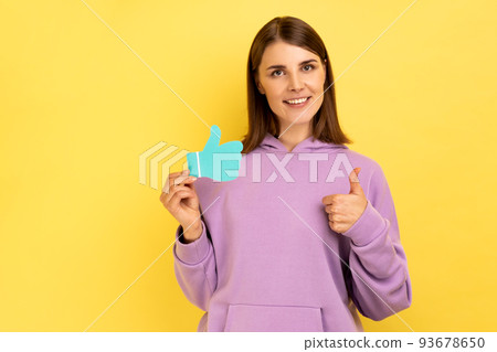 Smiling woman showing thumbs up blue paper sign, liking and recommending posts in social networks, asking to rate, wearing purple hoodie. Indoor studio shot isolated on yellow background. Smiling woman showing thumbs up blue paper sign, liking and recommending posts in social networks, asking to rate, wearing purple hoodie. Indoor studio shot isolated on yellow background. 93678650