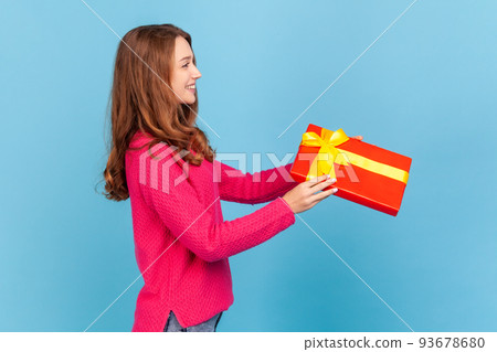 Side view portrait of open hearted woman wearing pink pullover, giving gift box and smiling broadly, congratulating on holiday and offering present. Indoor studio shot isolated on blue background. Side view portrait of open hearted woman wearing pink pullover, giving gift box and smiling broadly, congratulating on holiday and offering present. Indoor studio shot isolated on blue background. 93678680