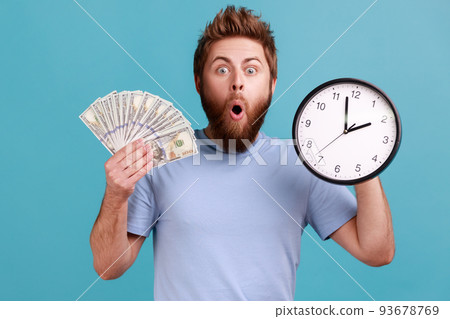 Portrait of surprised handsome bearded man holding big fan of dollar banknotes and wall clock, time is money, expressing astonishment. Indoor studio shot isolated on blue background. 93678769