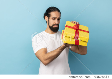 Portrait of unhappy man with beard wearing white T-shirt looking inside present box and expressing sadness, hoped to get another gift. Indoor studio shot isolated on blue background. 93678814