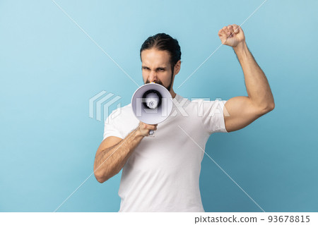 Portrait of angry man with beard wearing white T-shirt loudly screaming at megaphone, making announce, protesting, wants to be heard. Indoor studio shot isolated on blue background. 93678815