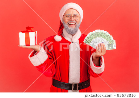 Good looking happy elderly man with gray beard wearing santa claus costume standing with Christmas present box and big amount of money in hands. Indoor studio shot isolated on red background. Good looking happy elderly man with gray beard wearing santa claus costume standing with Christmas present box and big amount of money in hands. Indoor studio shot isolated on red background. 93678816