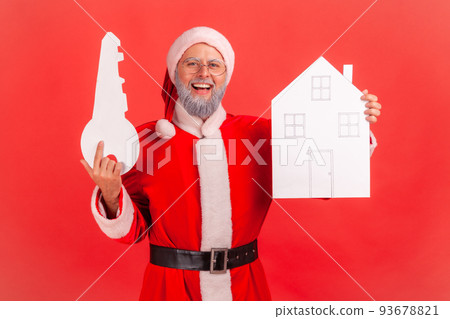 Smiling elderly man with gray beard wearing santa claus costume holding in hands paper house and key, being happy to buy new apartment. Indoor studio shot isolated on red background. 93678821