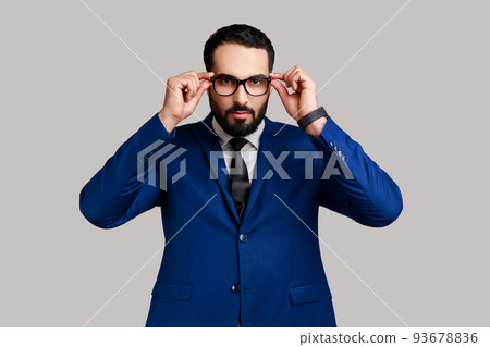 Portrait of serious bearded businessman putting off eyeglasses and looking at camera with bossy expression, wearing official style suit. Indoor studio shot isolated on gray background. Portrait of serious bearded businessman putting off eyeglasses and looking at camera with bossy expression, wearing official style suit. Indoor studio shot isolated on gray background. 93678836