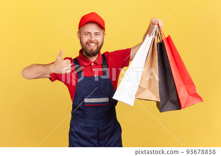 Portrait of happy smiling courier man in uniform holding shopping bags in his hand, showing thumb up, fast delivery from store, service industry. Indoor studio shot isolated on yellow background. Portrait of happy smiling courier man in uniform holding shopping bags in his hand, showing thumb up, fast delivery from store, service industry. Indoor studio shot isolated on yellow background. 93678838
