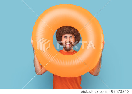 Portrait of man with Afro hairstyle with happy facial expression looking trough bright orange rubber ring, looking at camera, enjoying his vacation. Indoor studio shot isolated on blue background. 93678840