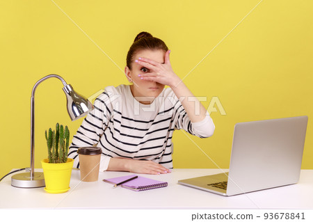 Curious woman manager sitting at workplace with laptop and spying, looking through fingers with interest, peeking secret gossip in office. Indoor studio studio shot isolated on yellow background. Curious woman manager sitting at workplace with laptop and spying, looking through fingers with interest, peeking secret gossip in office. Indoor studio studio shot isolated on yellow background. 93678841