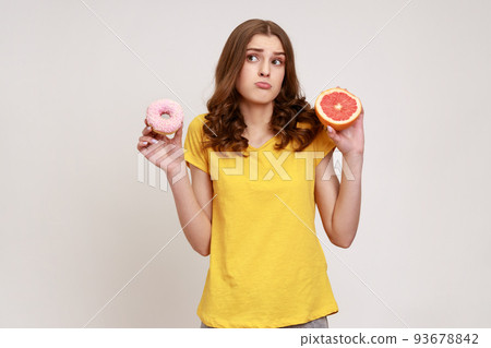 Healthy fruits vs high-calorie dessert. Portrait of confused curly-haired teen girl in casual style T-shirt choosing between grapefruit and donut. Indoor studio shot isolated on gray background. Healthy fruits vs high-calorie dessert. Portrait of confused curly-haired teen girl in casual style T-shirt choosing between grapefruit and donut. Indoor studio shot isolated on gray background. 93678842